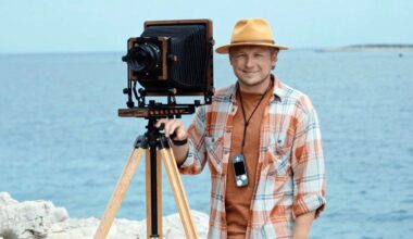 A man wearing a yellow hat and plaid shirt stands by the sea, smiling next to a vintage large-format camera mounted on a tripod. Rocky shore and calm blue water are in the background.