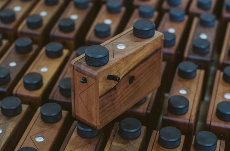 A close-up of a wooden camera with black dials, resting at an angle on top of many identical wooden cameras arranged in rows.