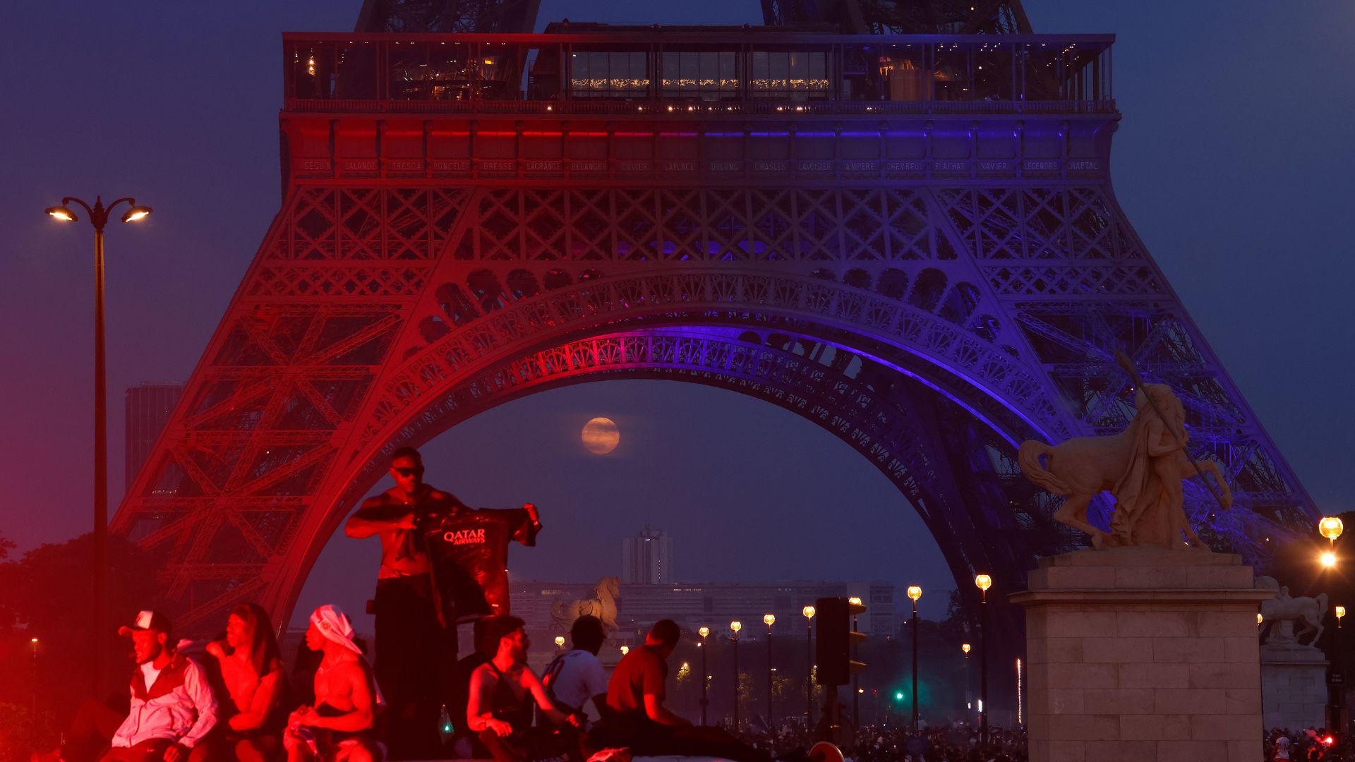 Fans celebrate as the full moon rises between the steel legs of the Eiffel Tower, which is lit in red and blue.