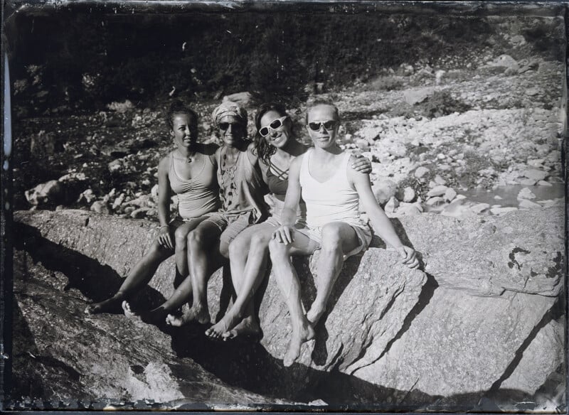 Four people sit side by side on a large rock outdoors, wearing casual summer clothes and sunglasses, smiling at the camera. Rocky terrain and trees are visible in the background. The image appears to be black and white.