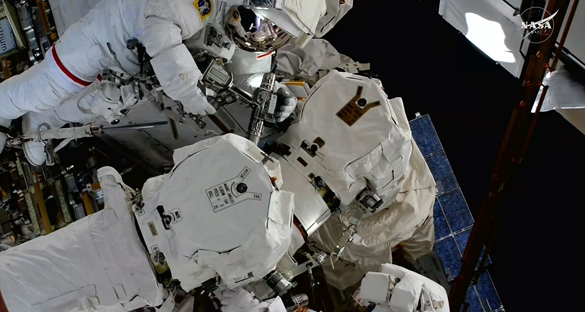 Two astronauts in white spacesuits work to repair a robotic arm outside of a space station