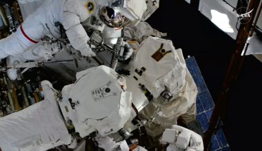 Two astronauts in white spacesuits work to repair a robotic arm outside of a space station