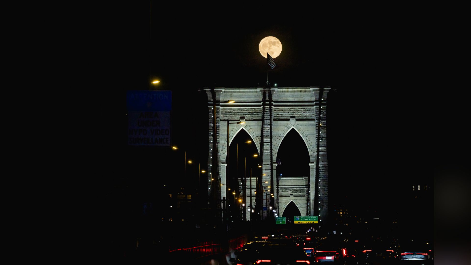 The full moon is photographed shining behind an American flag atop the Brroklyn Bridge, which is illuminated at night.