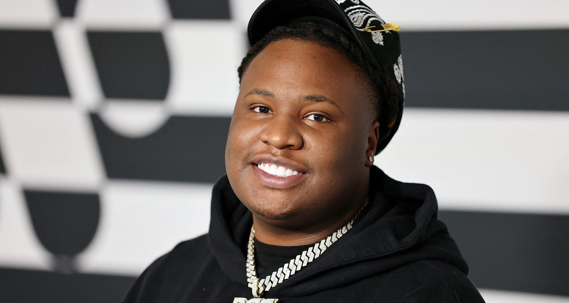A smiling person wearing a black hoodie and cap, with a chain necklace, stands against a checkered background.
