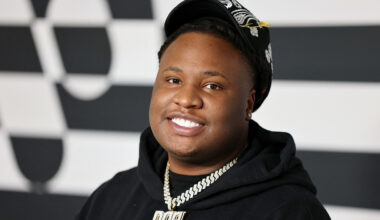 A smiling person wearing a black hoodie and cap, with a chain necklace, stands against a checkered background.