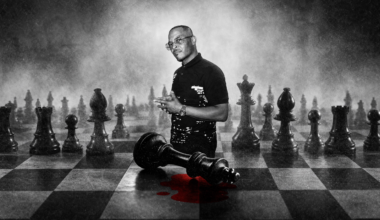 A man stands on a large chessboard with oversized pieces. A fallen king piece lies near him with a red stain beneath it.
