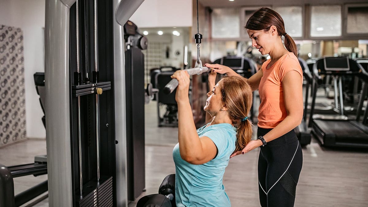 Senior woman doing lat pulldown exercise with trainer assistance in gym