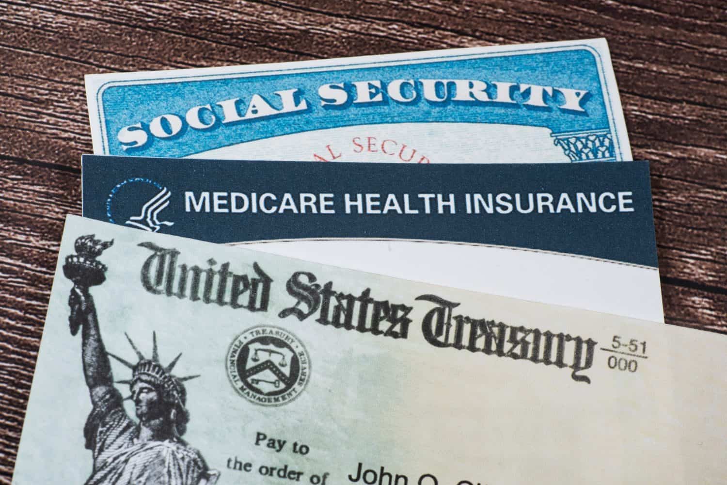 Three government-issued documents are stacked and partially overlapping on a dark wooden surface. The top document is a blue and white Social Security card, partially revealing the text 'SOCIAL SECURITY'. Below it is a dark blue and white Medicare Health Insurance card, displaying the text 'MEDICARE HEALTH INSURANCE' and the Medicare logo. At the bottom, partially visible, is a light green and white United States Treasury check, featuring a silhouette of the Statue of Liberty on the left and the words 'United States Treasury'.