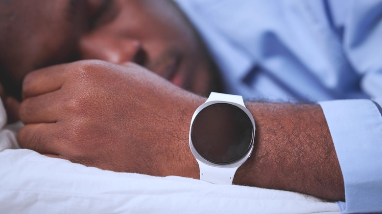 A man in a blue shirt laying in bed white white linens, a smartwatch with a white band is attached to their left wrist