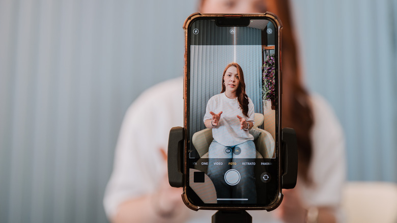 A woman sitting in front of a smartphone that is attached to a tripod and recording her