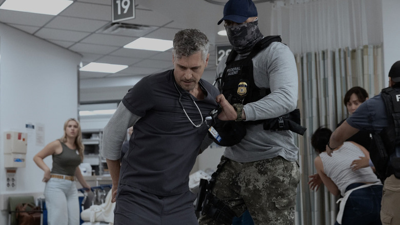A nurse in dark gray scrubs is arrested by a federal agent in a bullet proof vest wearing a face covering and a baseball cap.