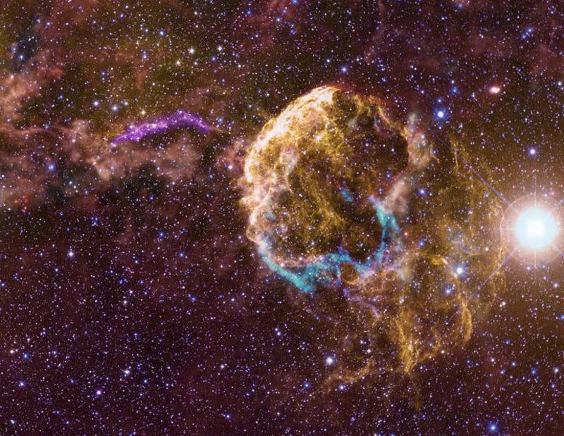 On a mottled reddish background speckled with stars, yellow threads of gas at center right trace out a dome-shaped structure from which curving tendrils extend toward the lower right. Blue and red filaments trace a ring around the dome’s base. A violet arc stretches right to left just above the center of the image, and irregular orange gas clouds become thicker toward the upper left.
