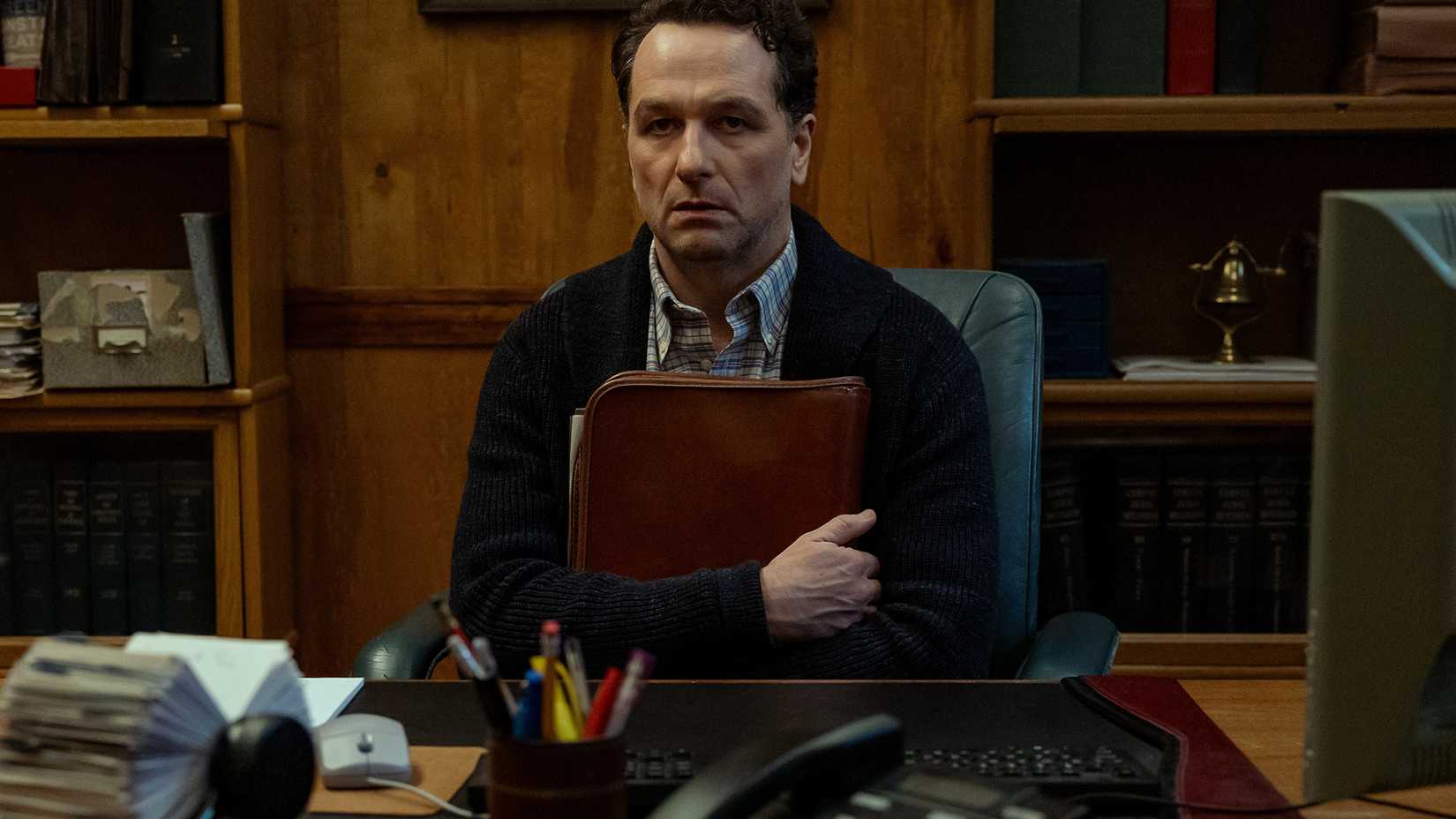 Tom sitting at his desk clutching a binder in Widow's Bay