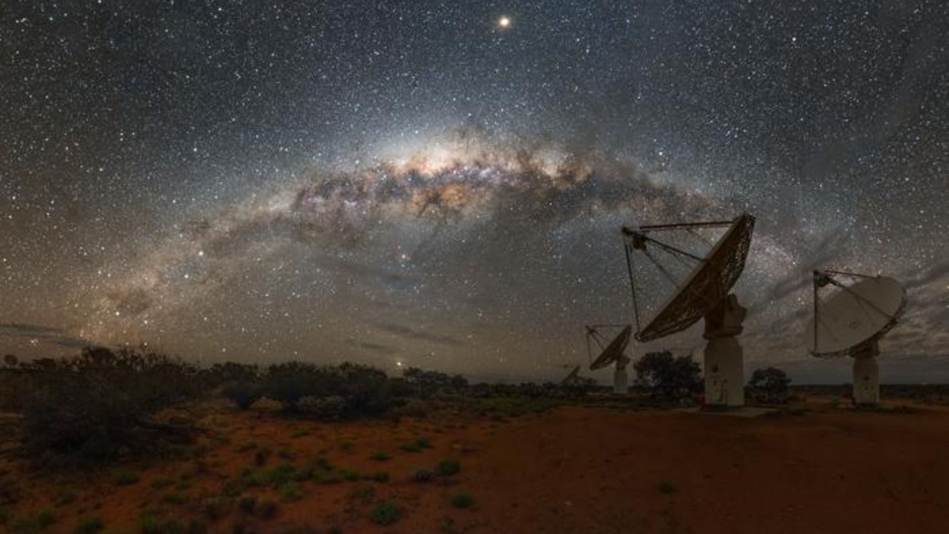 The Milky Way's heart glows beautifully in the sky. A bunch of radiowave detectors toward the right of the scene look up.