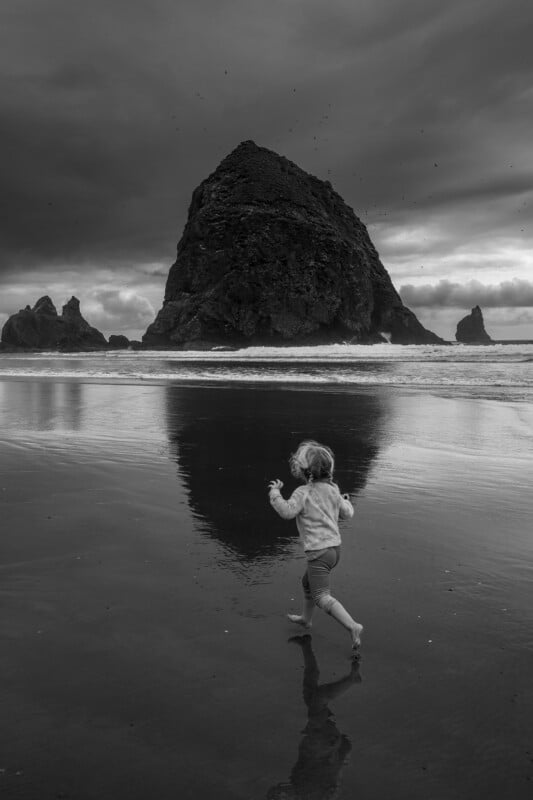 A small child runs barefoot on a wet beach toward a large, rocky sea stack under a dramatic, cloudy sky. The rock’s reflection is visible on the sand. The image is in black and white.