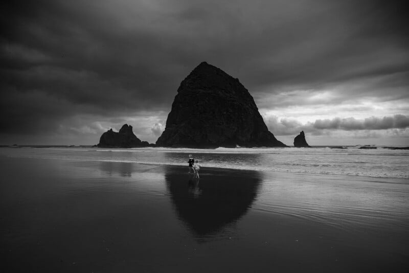 A person walks along a wet, reflective beach toward a large rock formation under dramatic, cloudy skies. The scene is in black and white, highlighting the moody atmosphere and natural landscape.