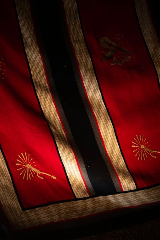 A red fabric with gold and black vertical stripes, embroidered with gold designs including a key and a sunburst pattern, partially illuminated by soft light.