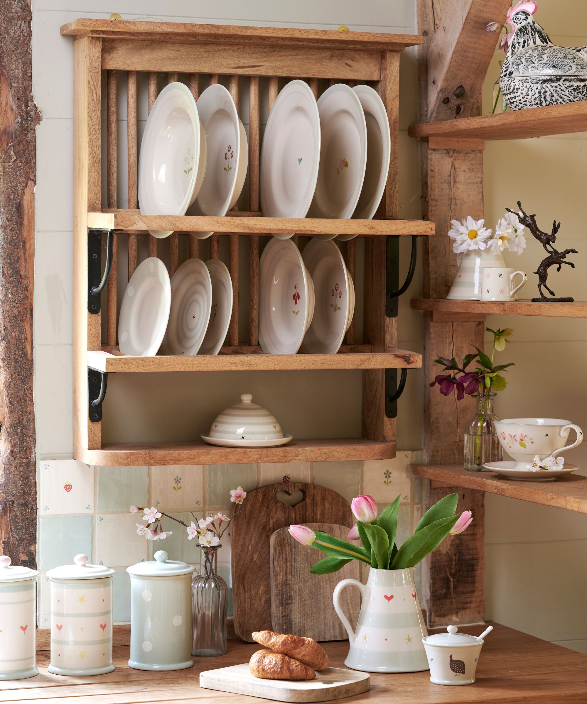 Fresh croissants on a wooden countertop next to a jug of pink tulips, below a wall-mounted wooden plate rack and pastel canisters