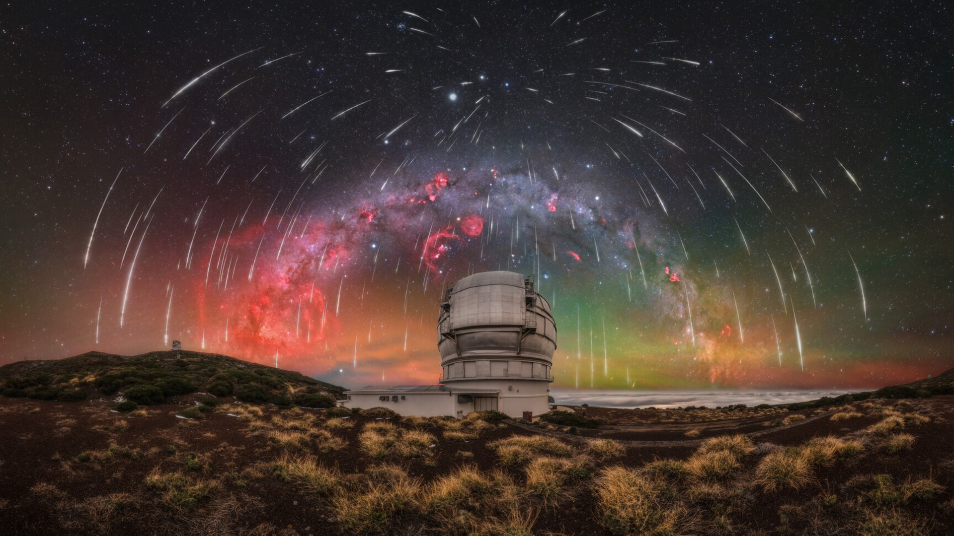 A long exposure image shows a glowing Milky Way galaxy with white streaks of meteors in the night sky over an observatory.