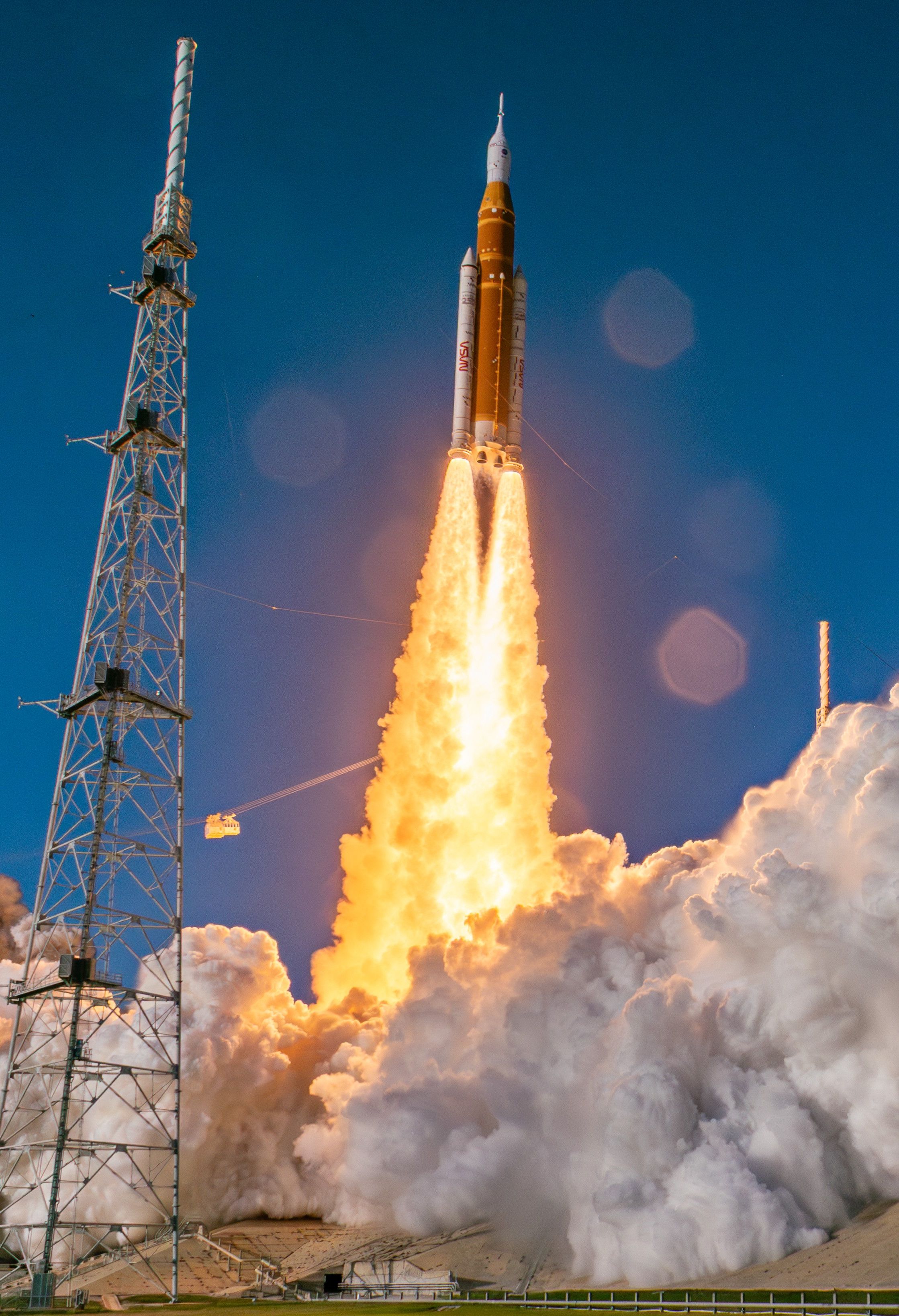 An orange rocket with two white side boosters blasts off into a blue sky.