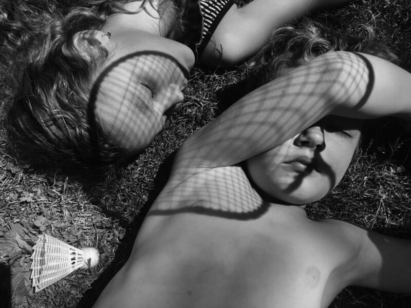 Two children lying on grass in sunlight, with one holding a badminton racket over their face, creating a shadow pattern. A shuttlecock is nearby. The image is in black and white.