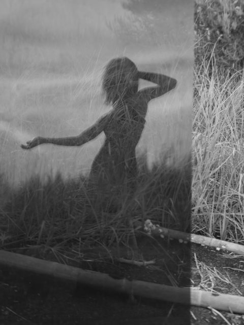A black and white photo shows the silhouette of a person posing amid tall grass, with the image partially obscured by a reflective or semi-transparent surface.
