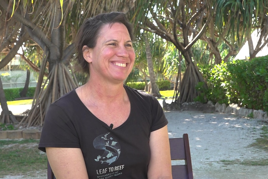 A woman with short brown hair smiling while sitting on a chair, a pathway behind her
