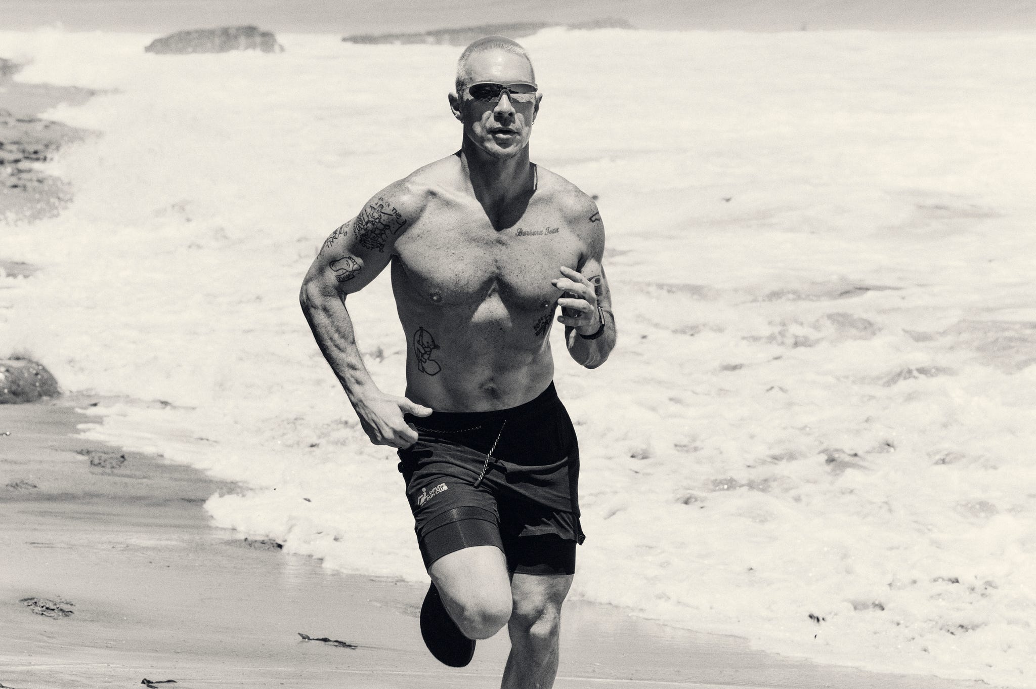 A man with tattoos runs along a beach with waves in the background.