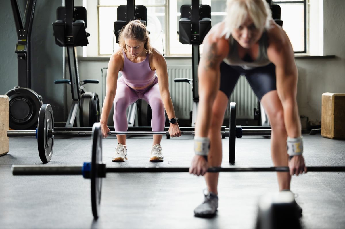 Two women position themselves over barbells, preparing for a deadlift movement. The image captures determination and proper form in a strength-focused workout.