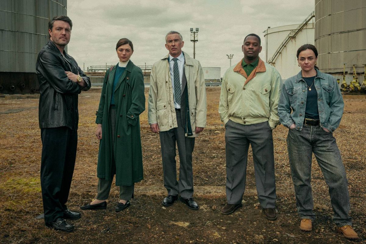 Five individuals, dressed in formal attire, are standing together in an open, dirt-covered area, with industrial structures visible in the background. They appear to be posing for a photograph.