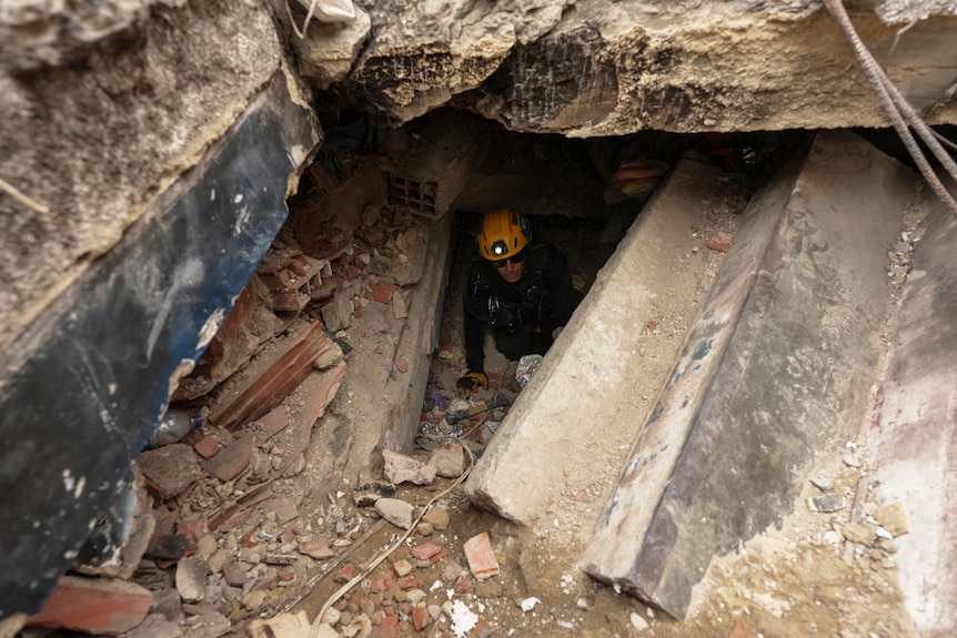 A man in a yellow search helmet crawls out from a mound of rubble.