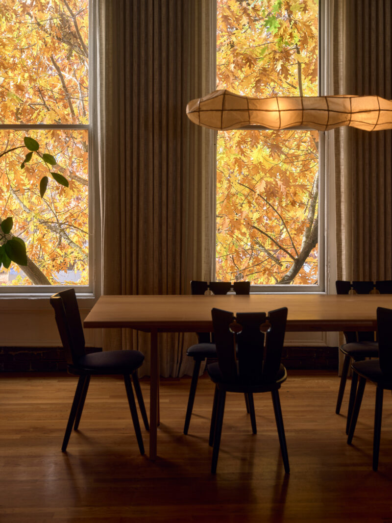 A wooden dining table with black chairs sits by large windows with autumn leaves visible outside; a modern light fixture hangs above.