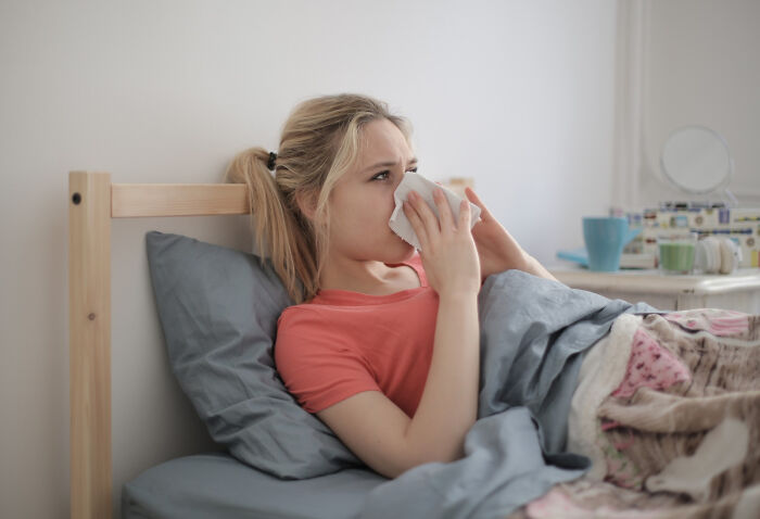 A young woman in bed, appearing sick, blowing her nose with a tissue, illustrating truths about the medical industry.
