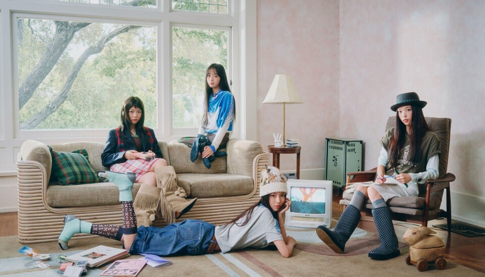 Several young adults sit and lie around a living room with a sofa, large windows and casual items on the floor.