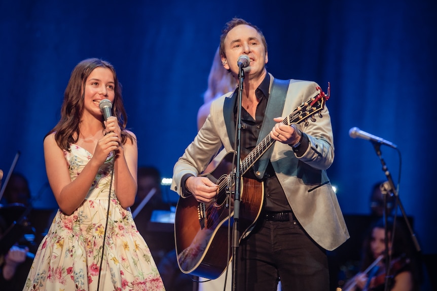 Damien performs on stage playing a guitar next to his daughter who wears a white floral dress.