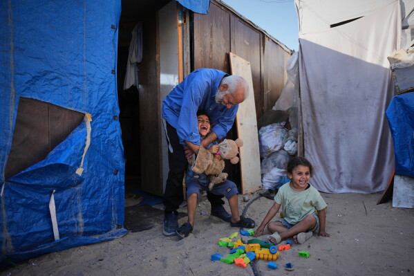 Mahmoud Nofal, 64, plays with his grandchildren, Tamim and Ranin Nofal, whom he has raised since their parents were killed in an Israeli airstrike, outside their tent in Khan Younis, southern Gaza Strip, June 2, 2026. (AP Photo/Abdel Kareem Hana)