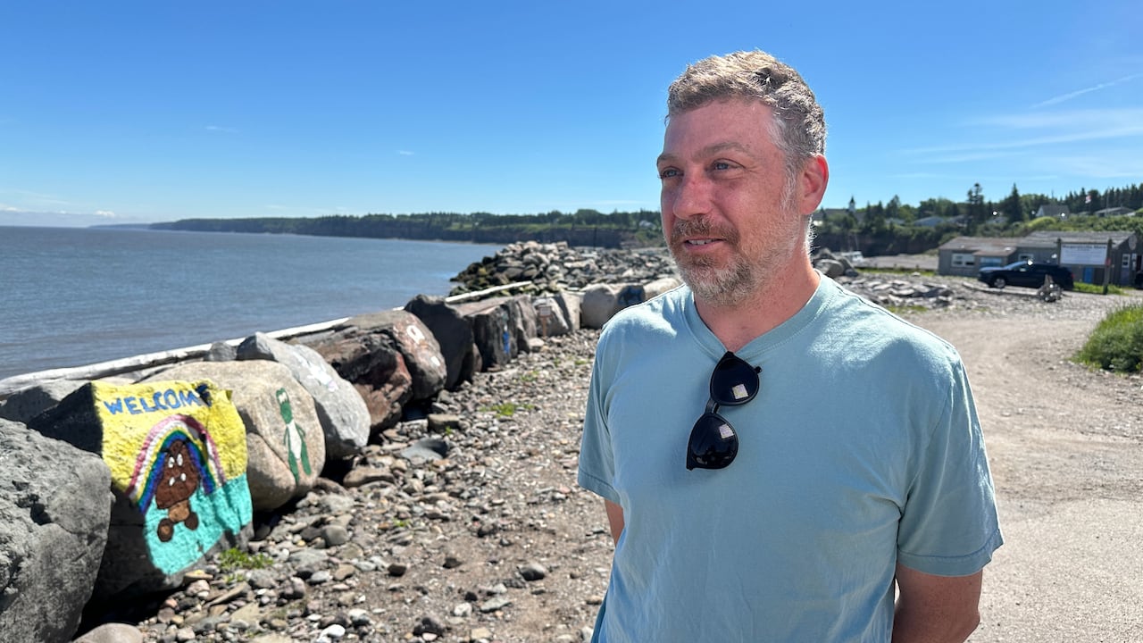A man stands between a gravel road and a line of large boulders next to a body of water.