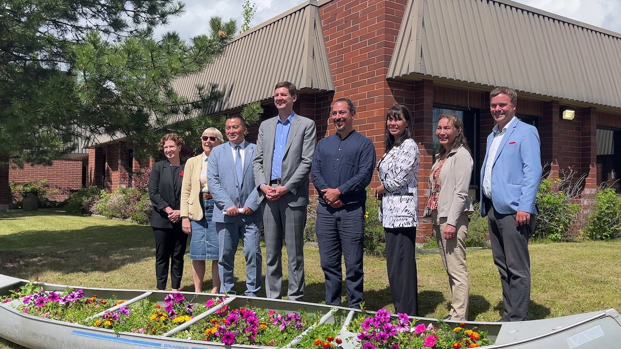 A group of eight people pose for a photo in front of a brick building and behind a canoe that is full of flowers.