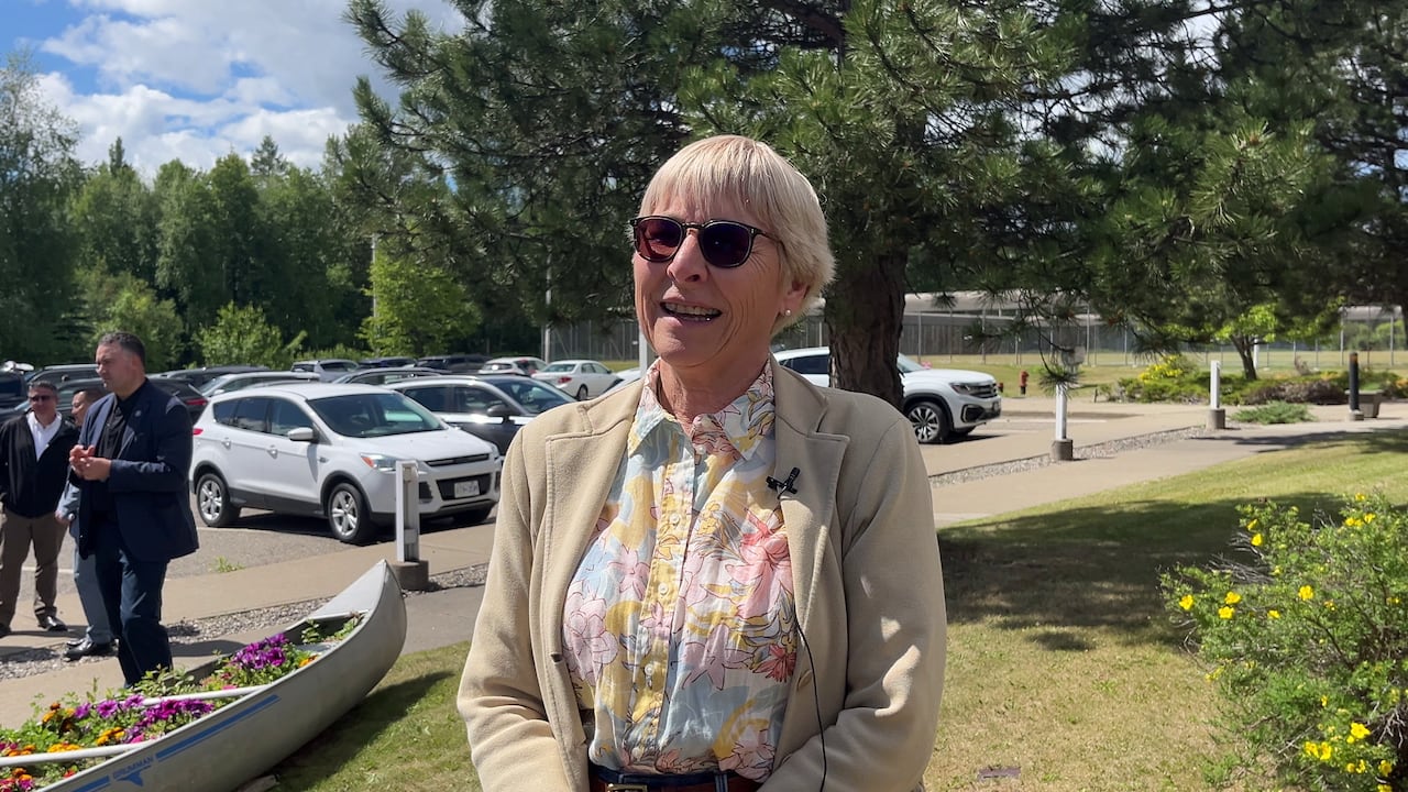 A woman with short hair and sun glasses stands in front of a tree on a sunny day.