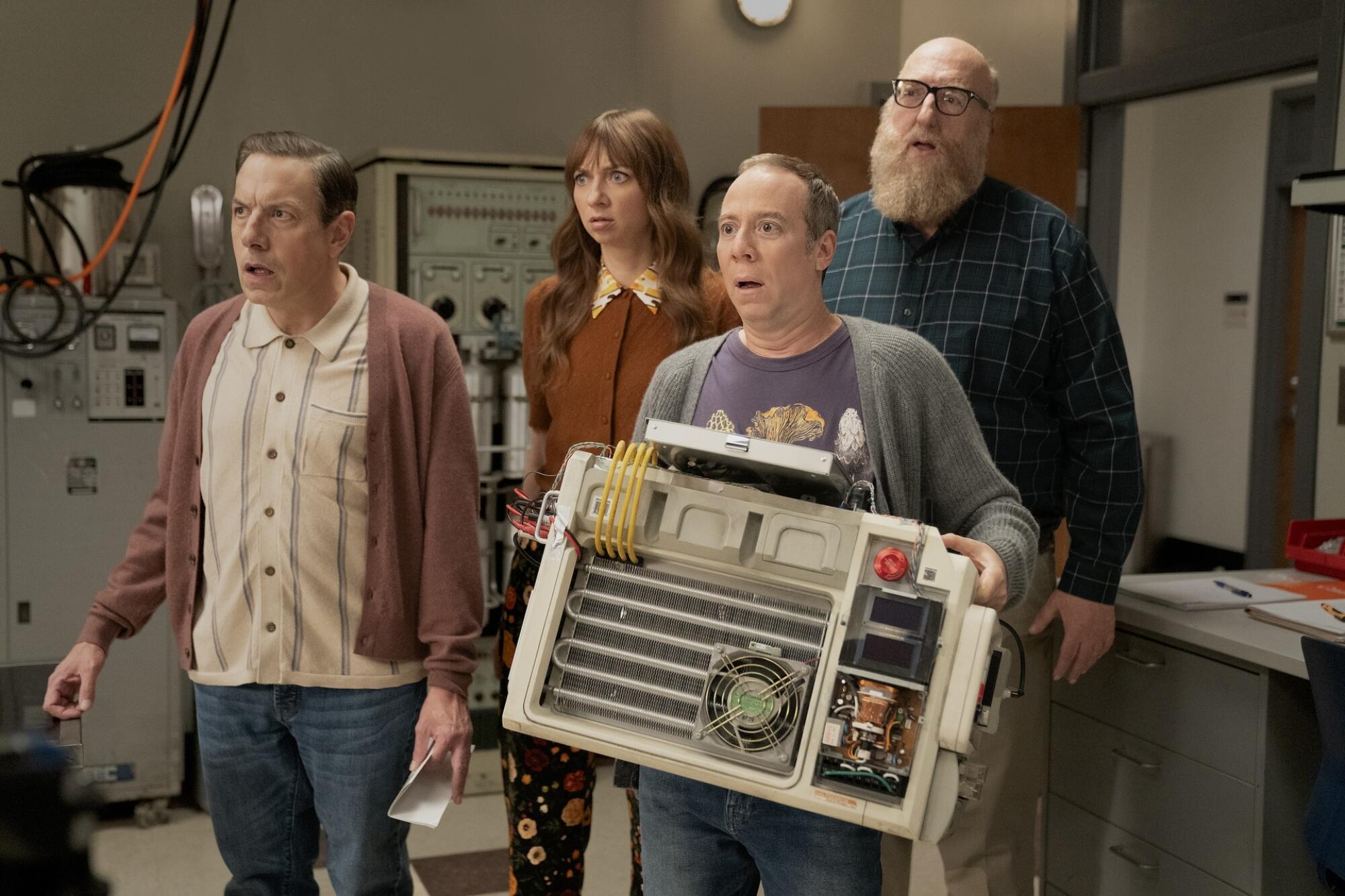 A man holding a big machine stands with a surprised face with two other men and a woman standing behind him.