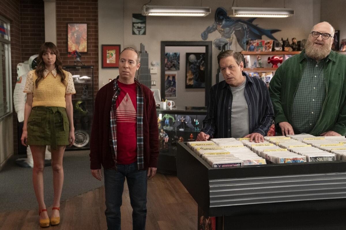 A woman stands in the background of three men standing near shelving filled with comic books.