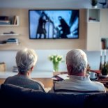 Rear view of two seniors with grey hair sitting on sofa in living room, looking television.