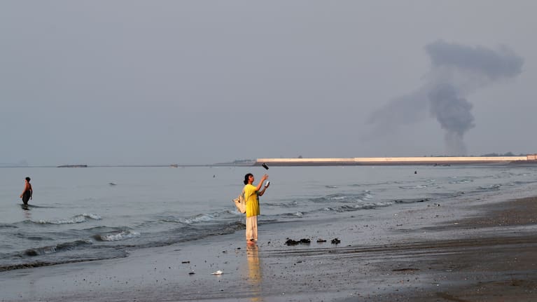 A woman stands at the water's edge along the Strait of Hormuz as a plume of smoke rises in the background following an explosion, off Bandar Abbas, Iran.