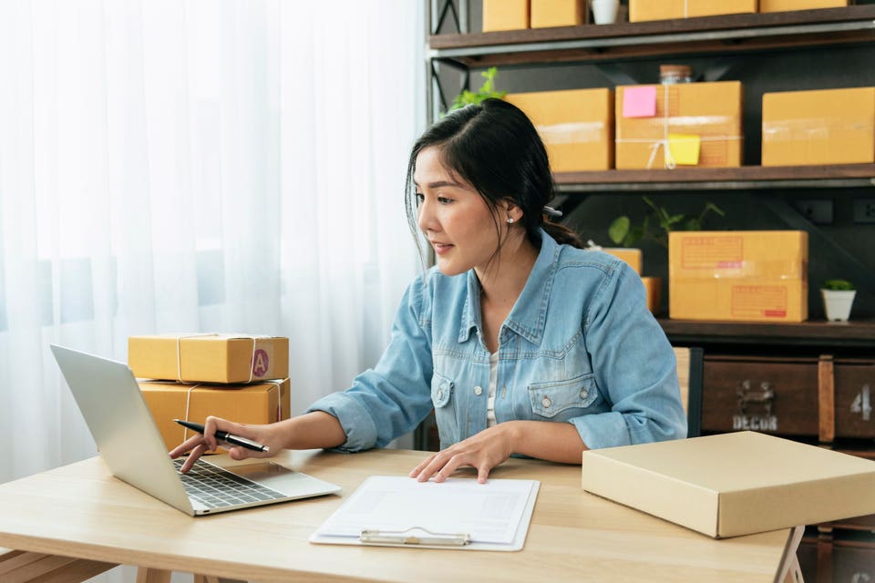 Woman types on laptop keyboard to check online order while preparing boxes for her commerce small business. Businesses with a $20,000 budget offer a larger upfront investment in equipment and inventory than a leaner budget, and provide more opportunity for marketing and growth initiatives.