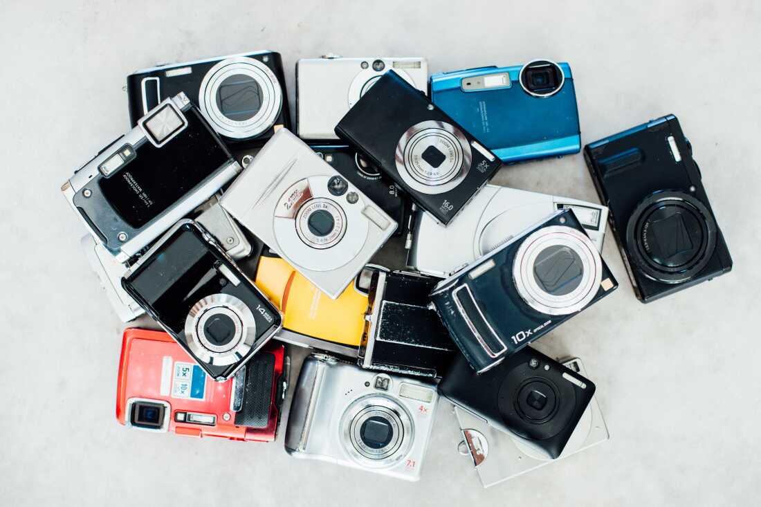 In this photo, about 20 digital cameras, all piled together in a jumble, are photographed from above. Most of the cameras are black or silver. One is red, another is blue and one is yellow.