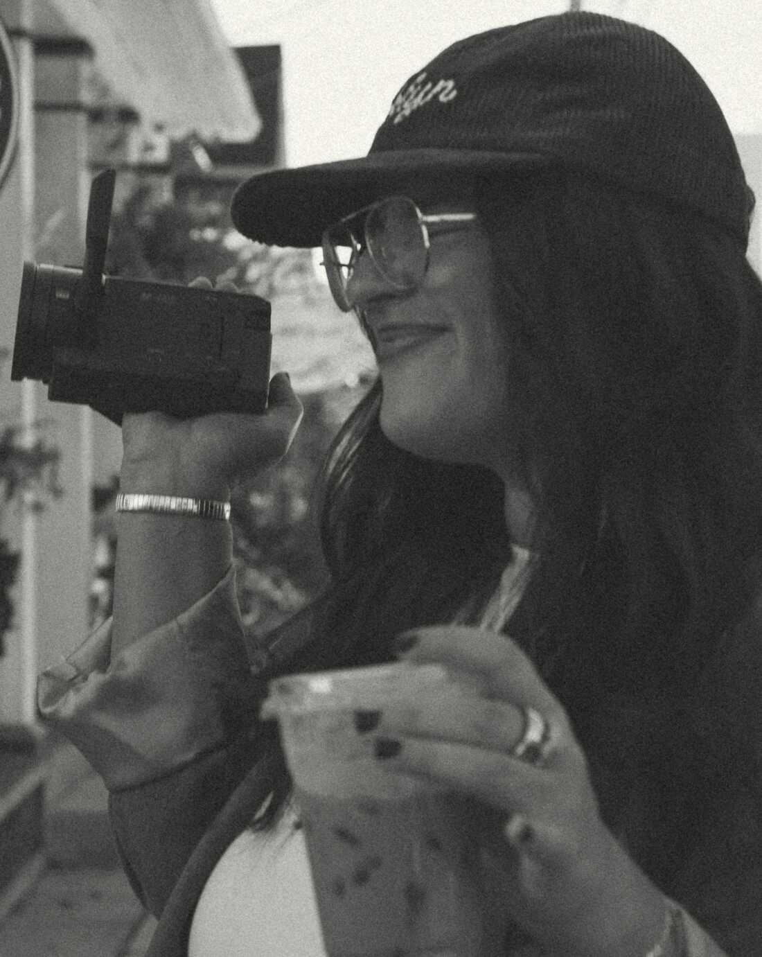 In this black-and-white photo, Christina Berkett is holding a camcorder in one hand and a drink in a to-go cup in the other. She's wearing a baseball cap and glasses, and she's smiling.