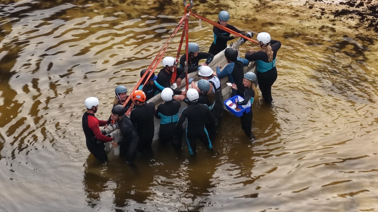 In a shot from above, people wearing helmets gather around a sling attached to orange straps.
