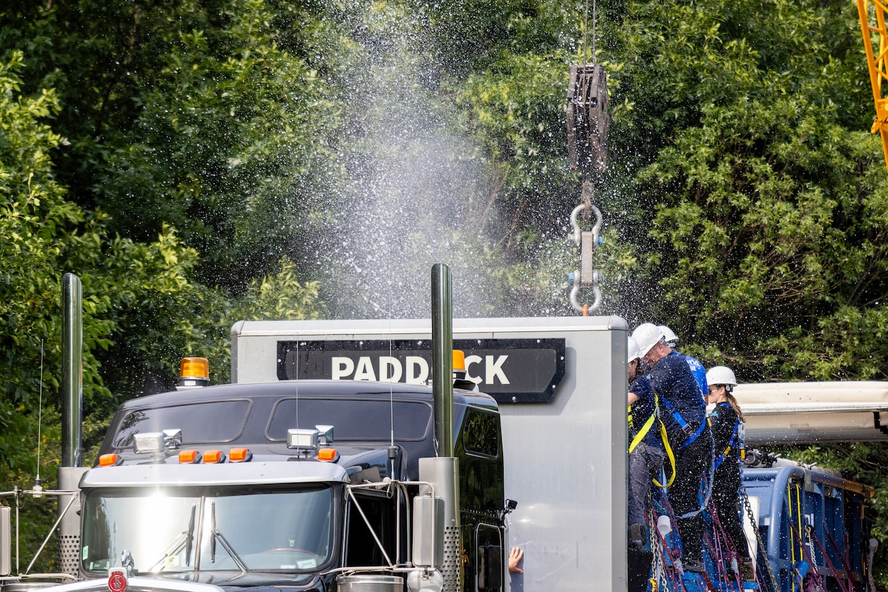 A water splashes into the sky behind transport truck hauling an open flatbed that says "PADDOCK". People in hard hats stand on the sides.