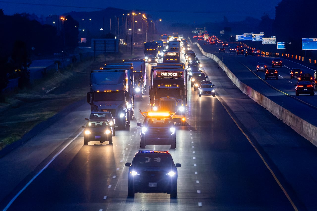 Transport trucks move along a multilane highway at night. Two of them say "PADDOCK" in illuminated letters.
