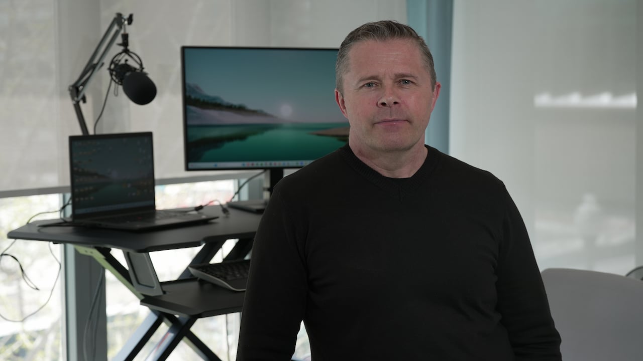 A man in a black shirt poses for a photo. There is a desk and a computer behind him.